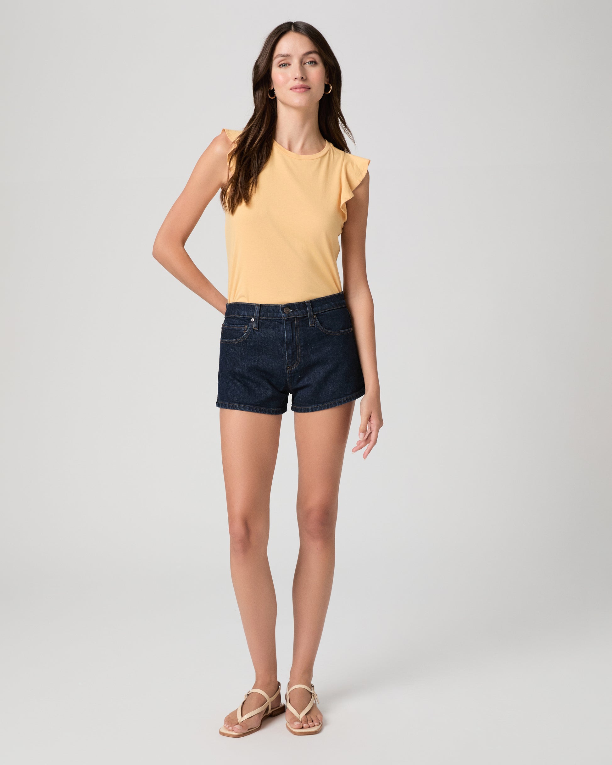 Woman wearing a yellow sleeveless top and denim shorts on a plain background