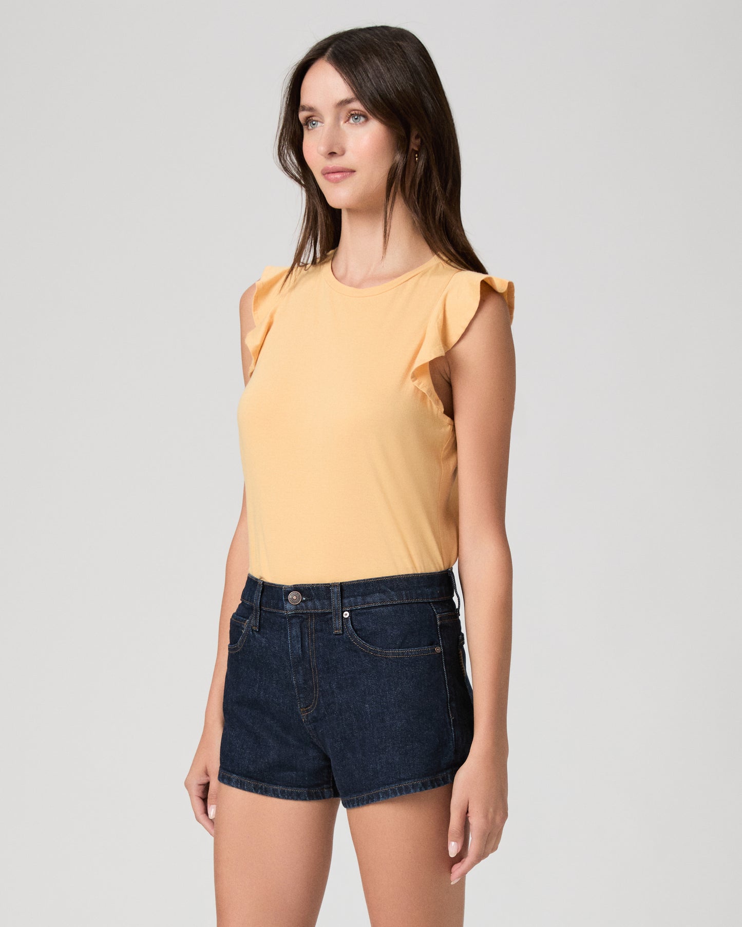 Woman wearing a yellow sleeveless top and denim shorts on a plain background