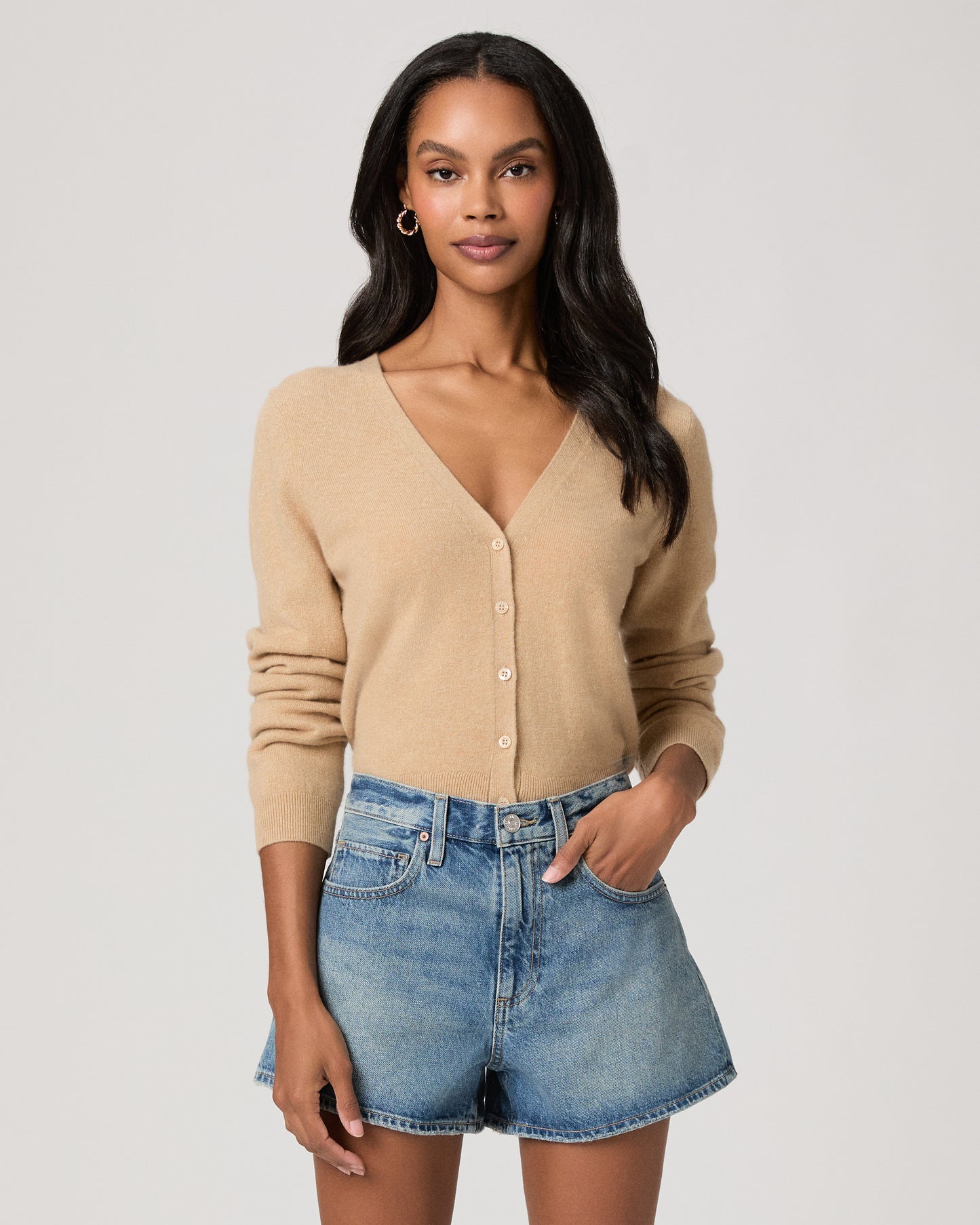 Woman wearing denim shorts and cream color long sleeve top on a plain background