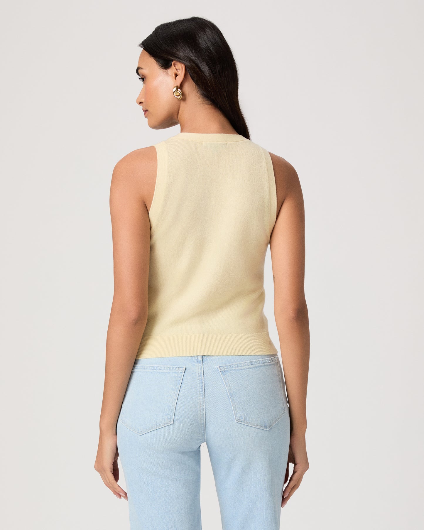 Woman wearing a light yellow sleeveless top and light blue jeans on a plain background