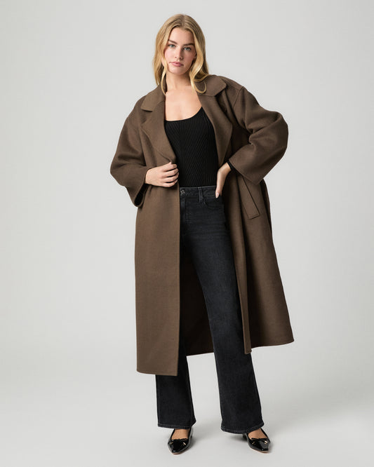 Full-length view of model with one hand on her hip, wearing a long brown wool coat over a black top with the Sofia 31 Inch Flare Jean in Solstice Black and black flats