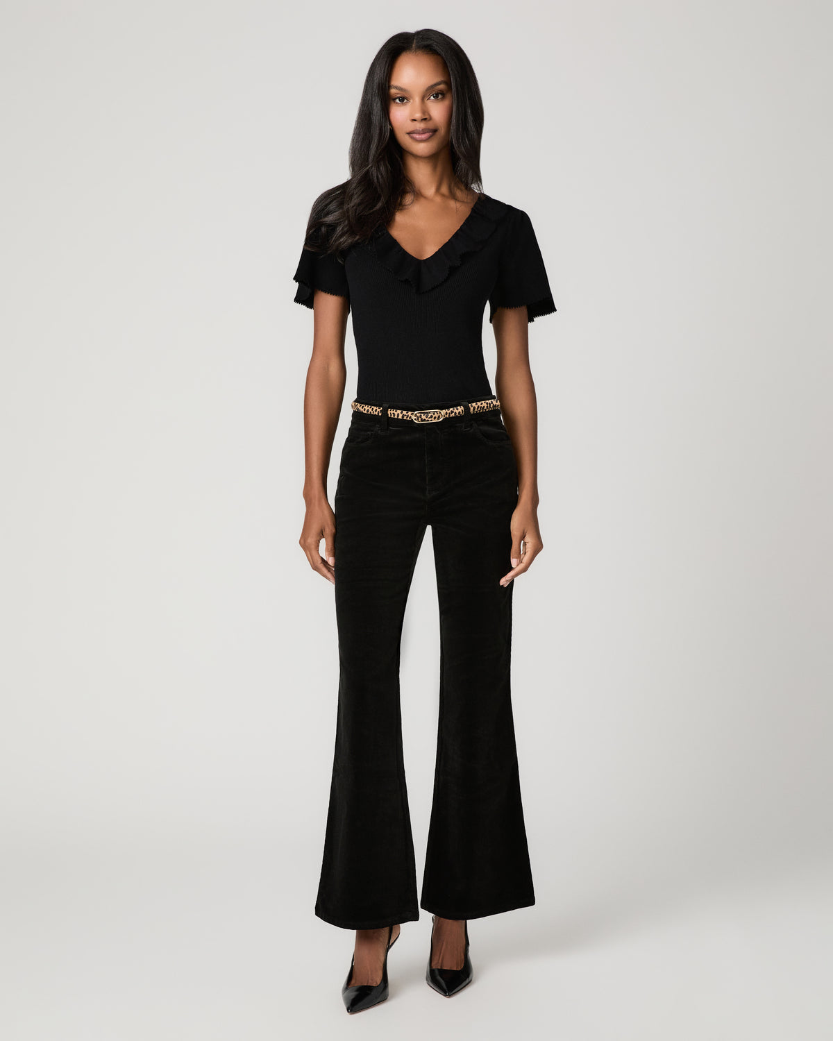 Full-length front view of a woman wearing a black short-sleeve V-neck top with a thin belt and black corduroy flare pants, paired with black pointed-toe heels.
