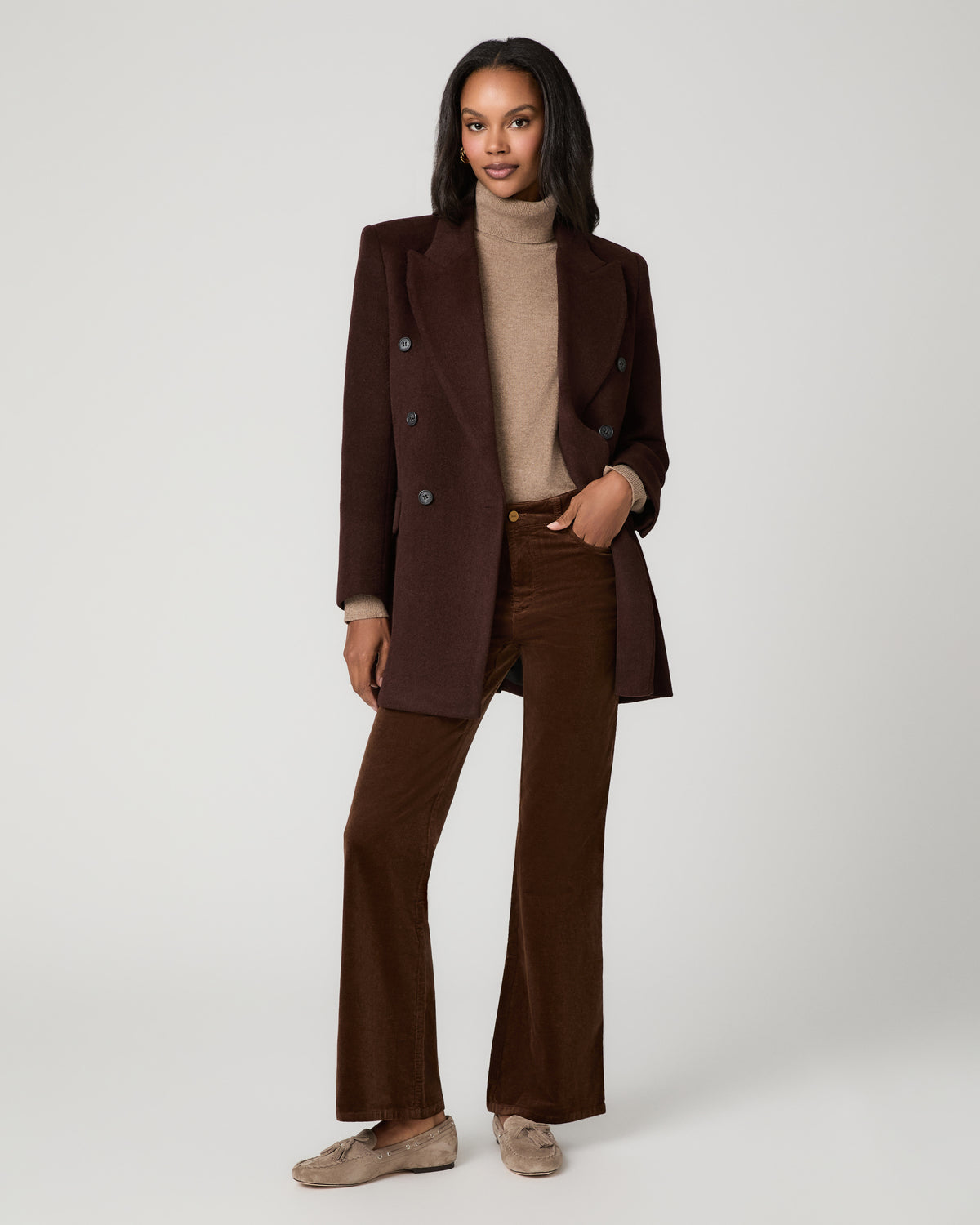 Full-length front view of a woman wearing a brown blazer over a tan turtleneck sweater and hickory brown corduroy flare pants, with one hand in her pocket, paired with tan flats.