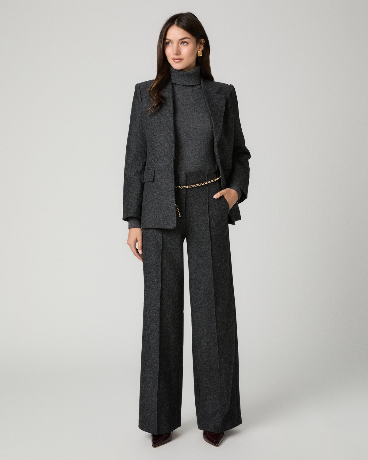 A model stands against a light gray studio background wearing a dark gray tailored suit set consisting of a turtleneck top, matching blazer, and wide-leg trousers, styled with a thin gold chain belt.