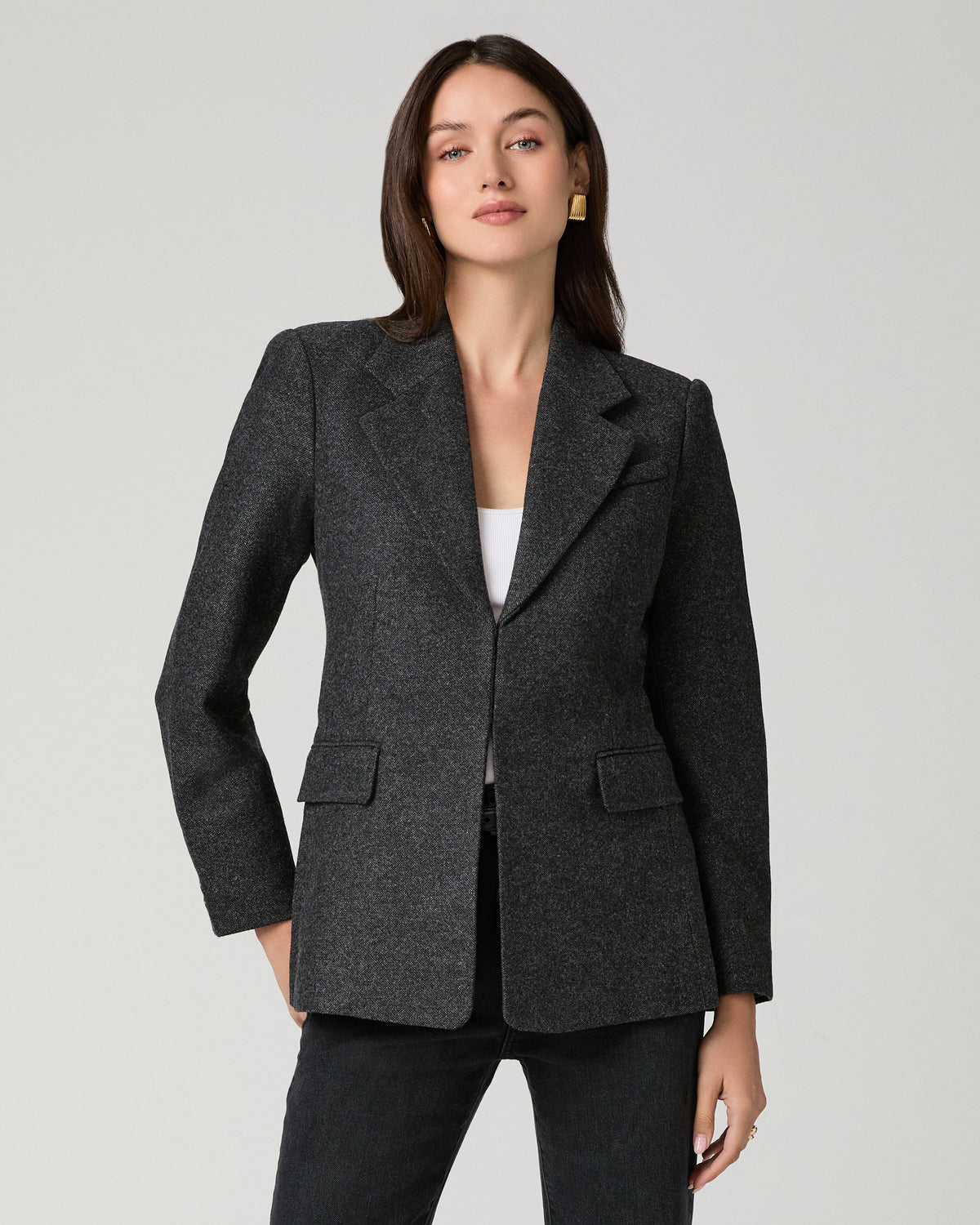 A woman modeling a dark charcoal gray textured blazer with notch lapels and flap pockets, worn over a white top and black pants, against a light gray studio background.