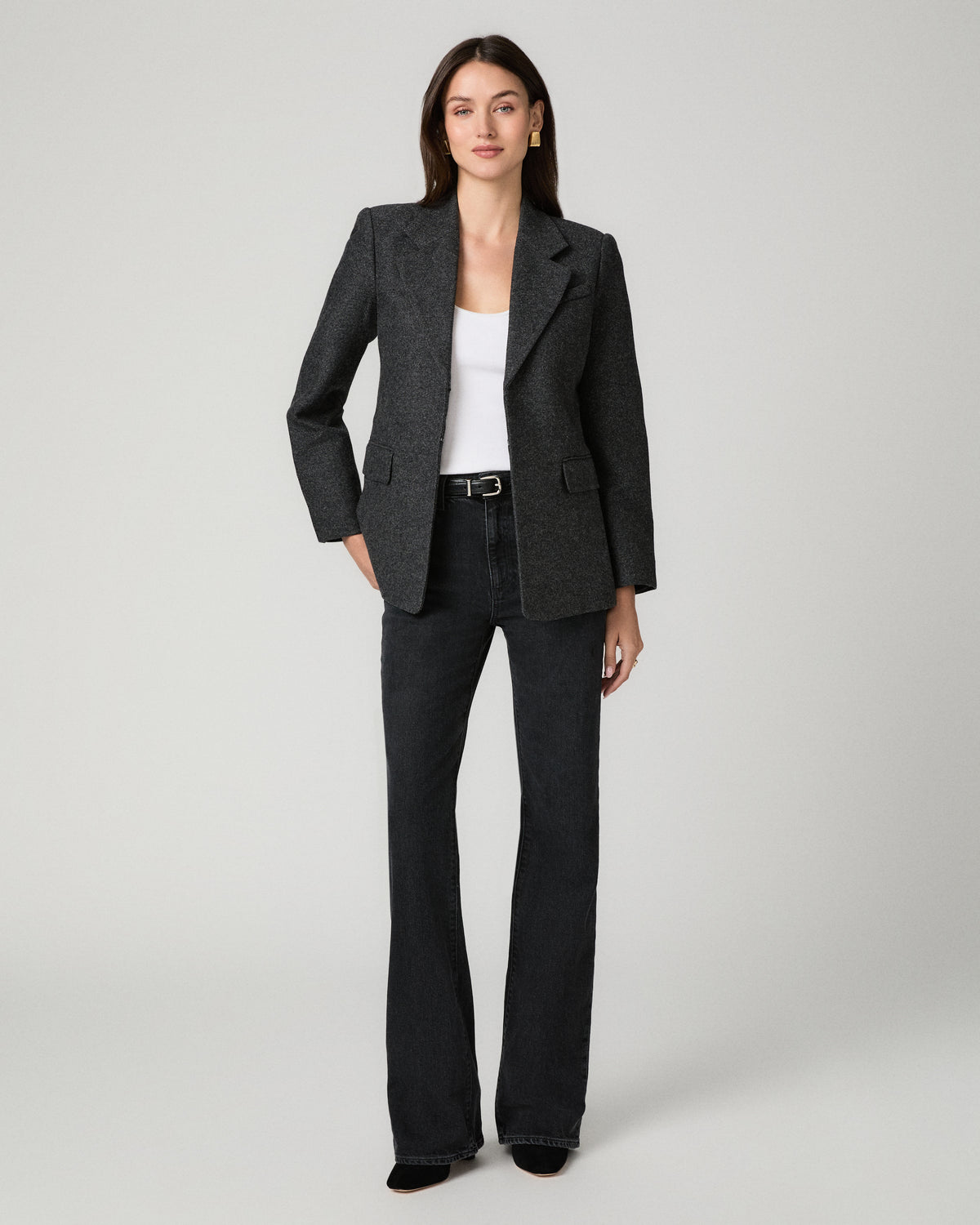 A woman modeling a dark gray textured blazer with dark charcoal flare jeans and a white tank top, standing against a light gray studio background.
