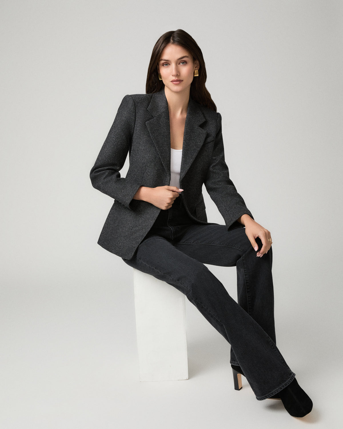 A woman models a dark gray textured blazer and matching wide-leg trousers with a white top and black heels, seated on a white block against a light gray studio background.