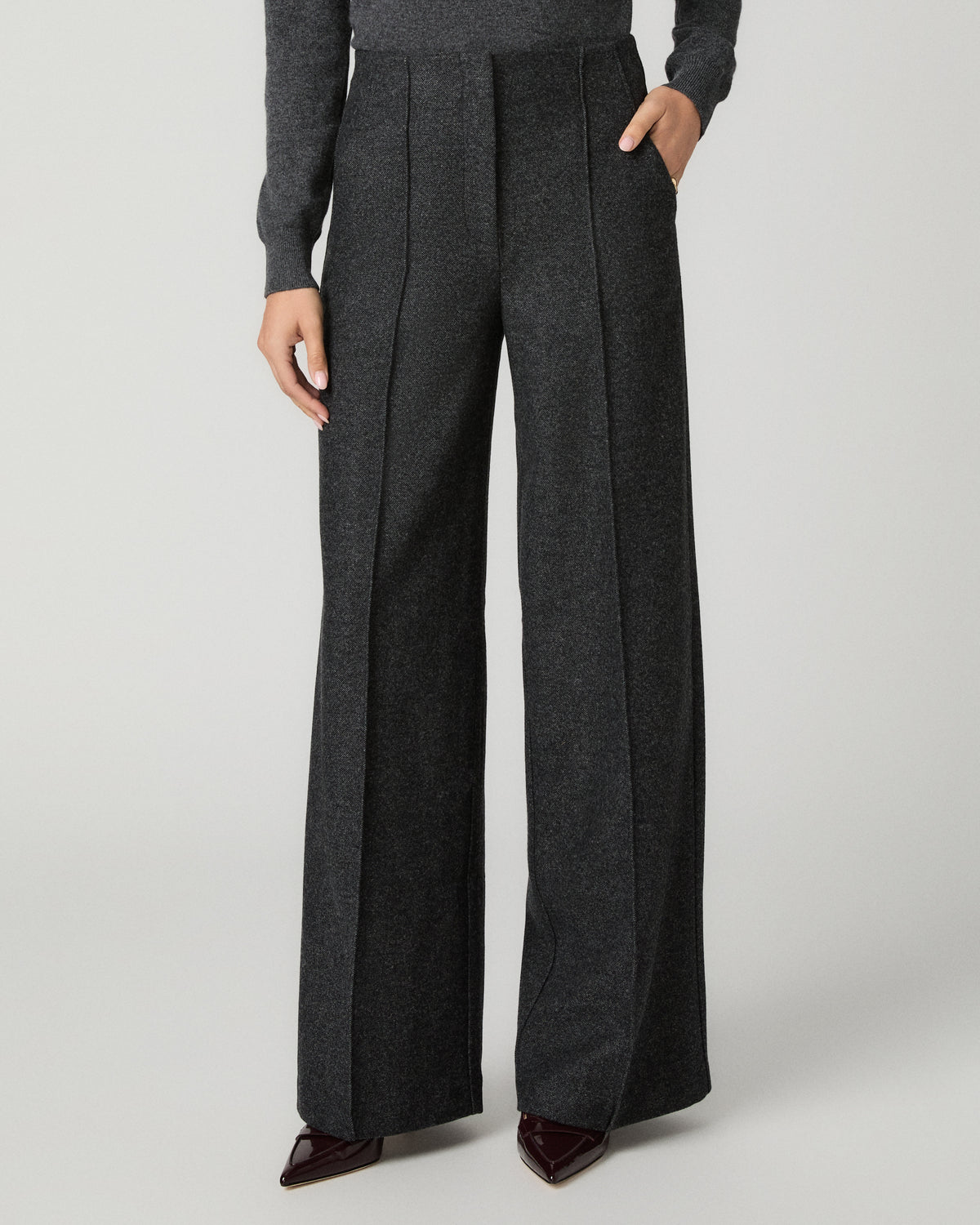 A model wearing dark gray wide-leg pleated trousers with a matching gray long-sleeve top and dark brown pointed-toe heels, shown from the waist down on a light gray studio background.