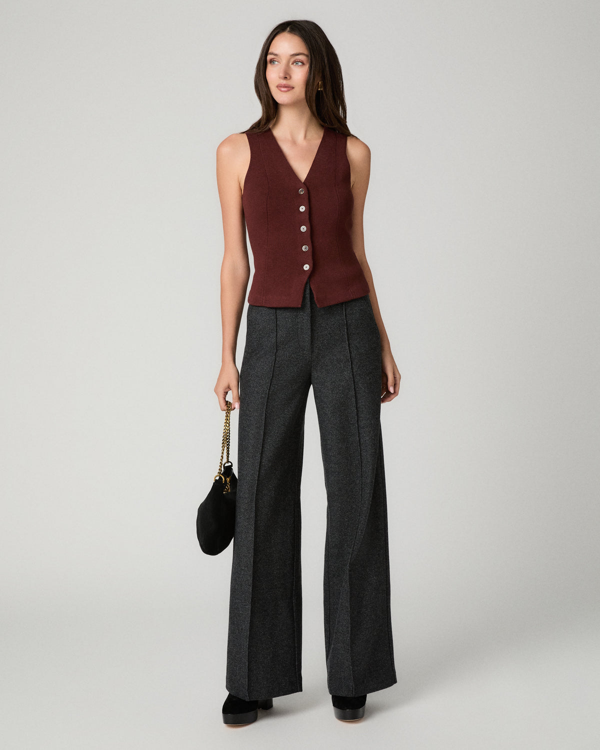 A model stands against a light gray studio background wearing a dark charcoal gray tailored set consisting of a structured turtleneck blazer and matching wide-leg trousers, styled with a thin gold chain belt and dark pointed shoes.