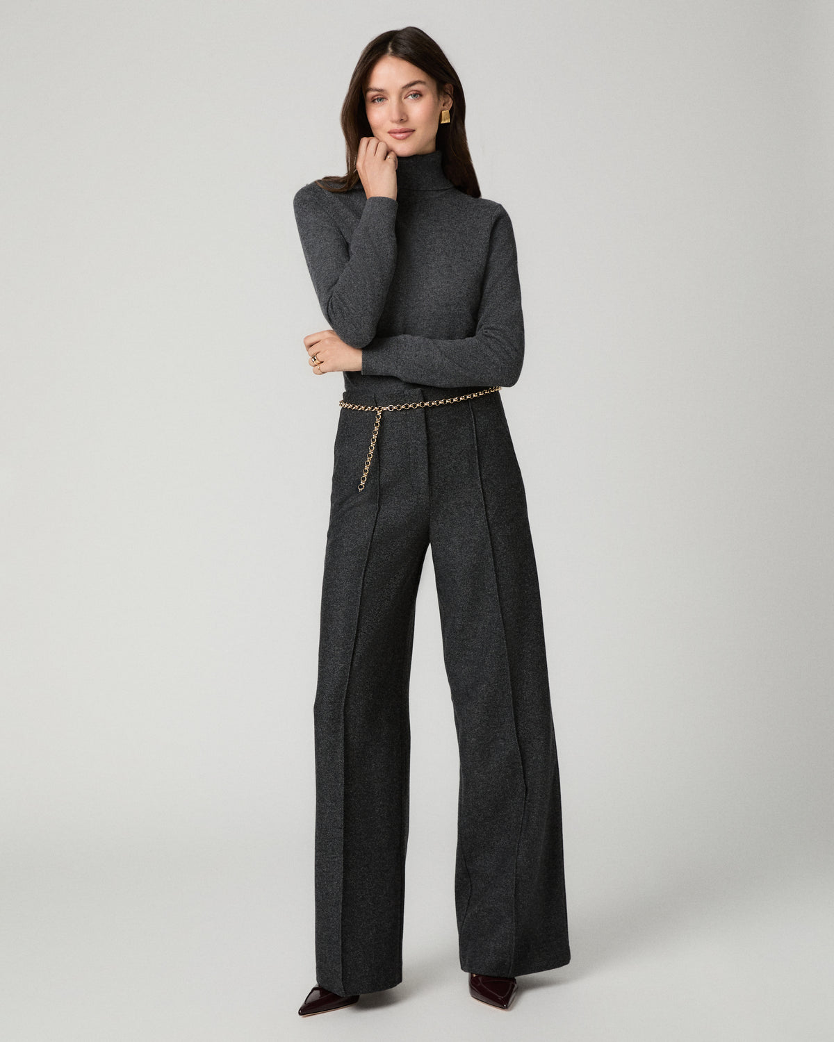 A model stands against a light gray studio background wearing a dark charcoal gray tailored set consisting of a structured turtleneck blazer and matching wide-leg trousers, styled with a thin gold chain belt and dark pointed shoes.