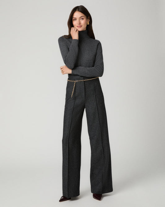 A model stands against a light gray studio background wearing a dark charcoal gray tailored set consisting of a structured turtleneck blazer and matching wide-leg trousers, styled with a thin gold chain belt and dark pointed shoes.