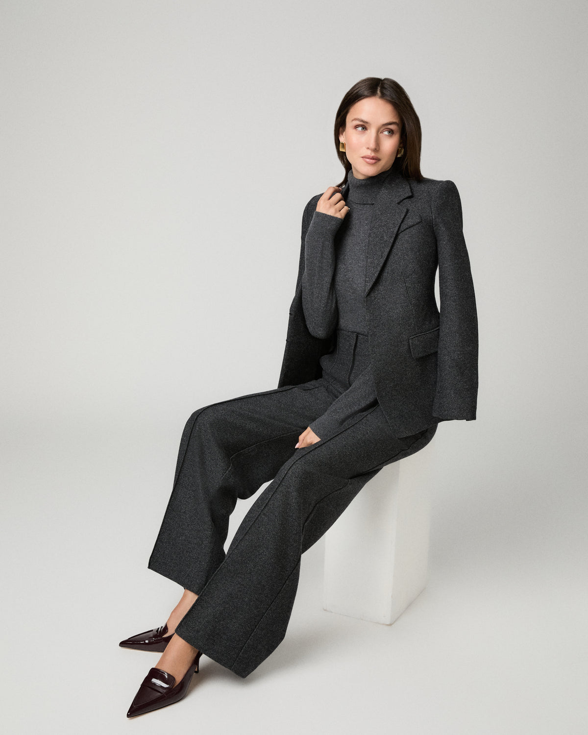 A model wearing a dark gray tailored suit with a structured blazer, matching trousers, and a turtleneck sweater, seated on a white block against a light gray studio background.