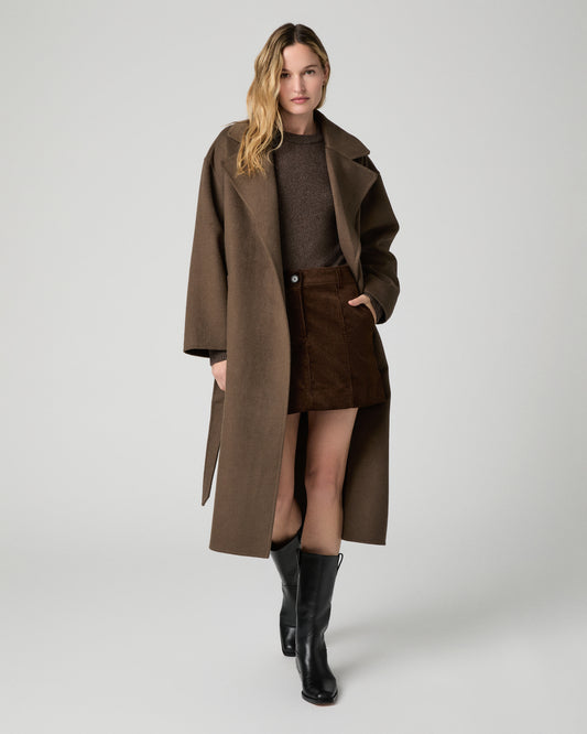 Women wearing a brown coat over a brown sweater and brown skirt, paired with black boots against a white background