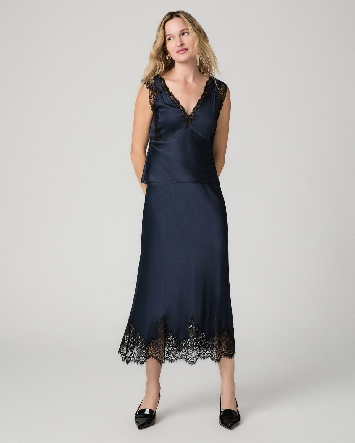 Full-length front view of a woman wearing the navy satin Kelso Skirt with a lace hem, paired with a matching navy V-neck top and black heels.