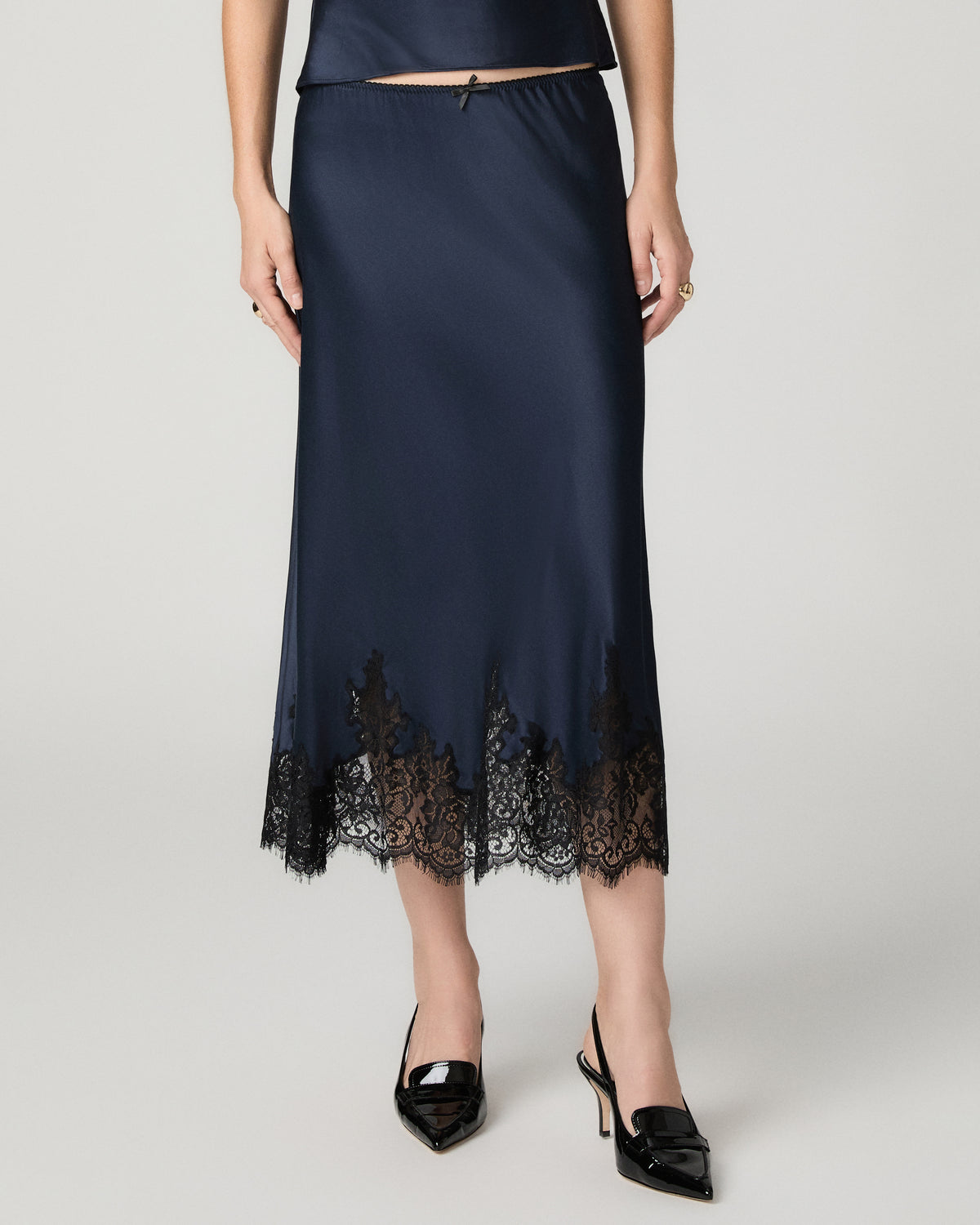 Close-up front view of the navy satin Kelso Skirt's lace hem, paired with black pointed heels.