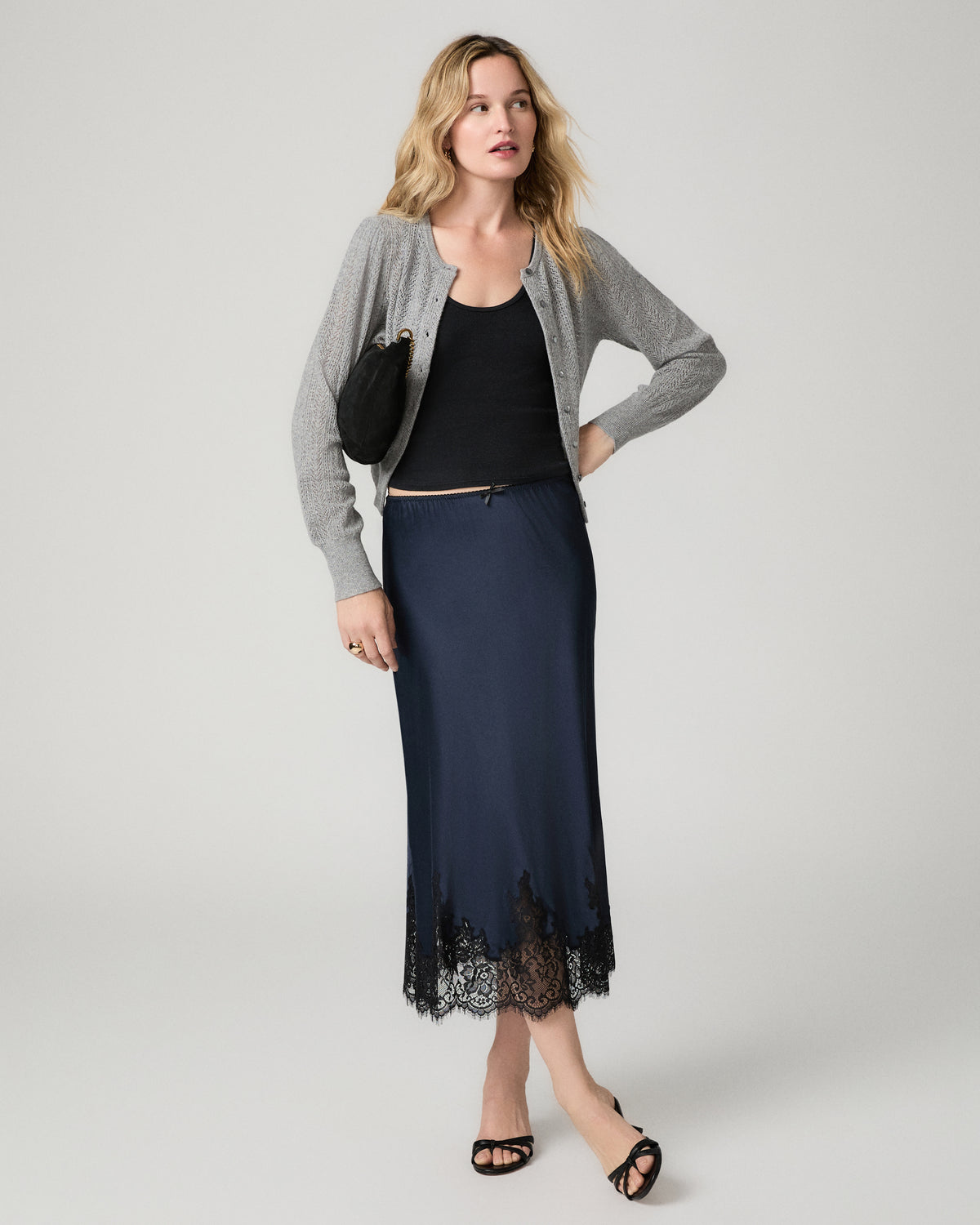 Full-length front view of a woman wearing the navy satin Kelso Skirt with a black top and gray cardigan, holding a small black clutch.