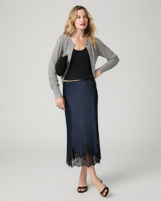 Full-length front view of a woman wearing the navy satin Kelso Skirt with a black top and gray cardigan, holding a small black clutch.