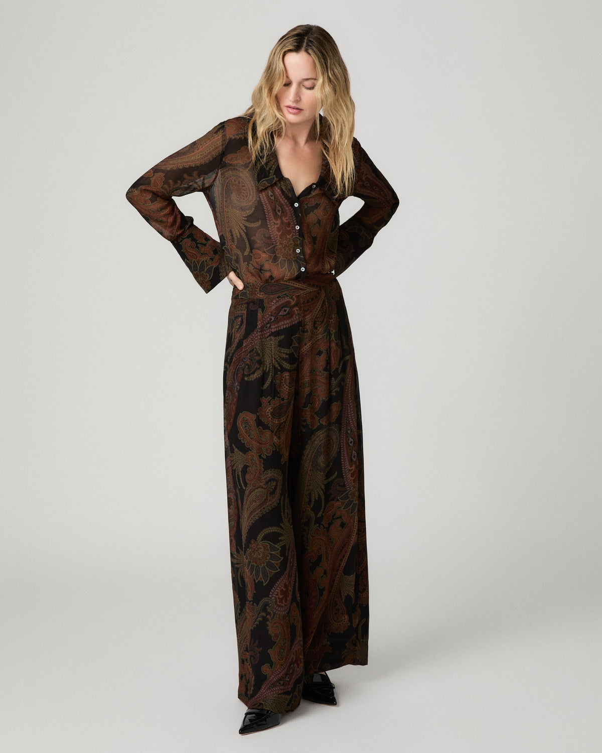 Full-length front view of a woman wearing the black and brown paisley-print Rosalinda Blouse tucked into matching wide-leg paisley pants, one hand on her hip.