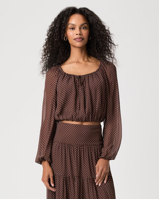 A woman modeling a brown long-sleeve brown polka dot blouse with a matching midi skirt and brown sandals against a light white background.