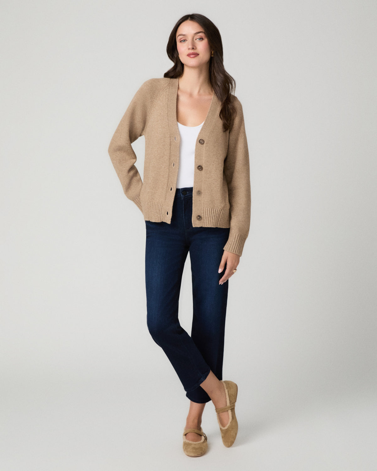 A woman modeling a beige knit cardigan with brown buttons, worn over a white top with dark blue jeans and tan flats, photographed against a light gray studio background.
