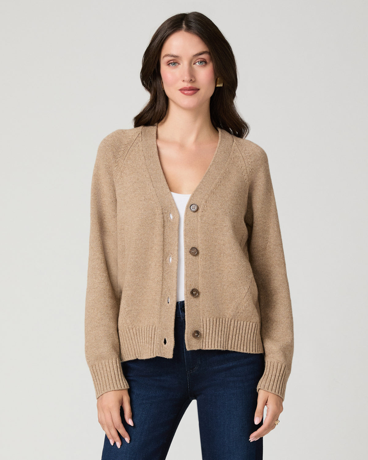 A woman modeling a beige knit cardigan with brown buttons, worn over a white top and dark blue jeans, against a light gray studio background.