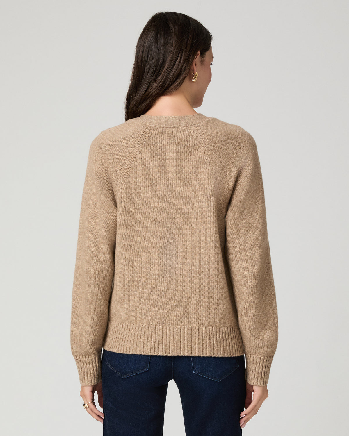 A woman shown from the back wearing a beige knit sweater and dark blue jeans against a light gray studio background.