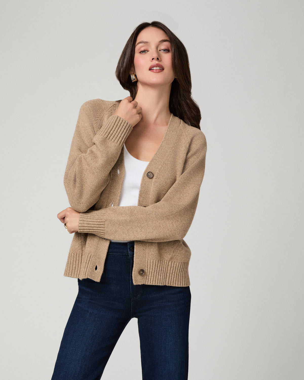 A woman modeling a beige knit cardigan with dark buttons, worn over a white top and dark blue jeans, against a light gray studio background.