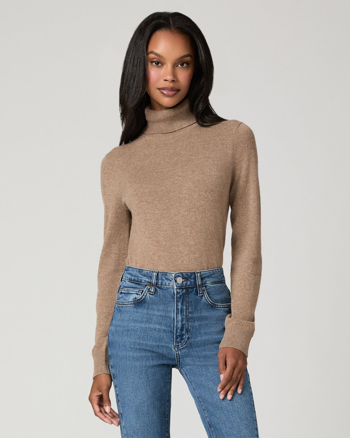 A woman modeling a beige ribbed turtleneck sweater paired with blue jeans, photographed against a solid light gray studio background.