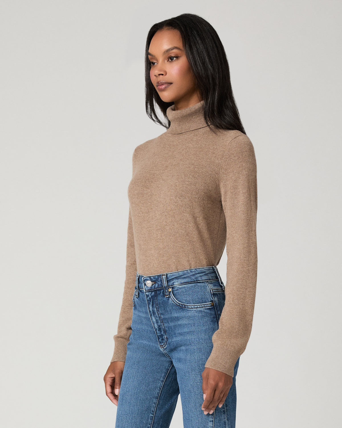 A model stands against a light gray studio background wearing a beige ribbed turtleneck sweater and blue high-waisted flare jeans with decorative silver buttons down the outer leg seams.