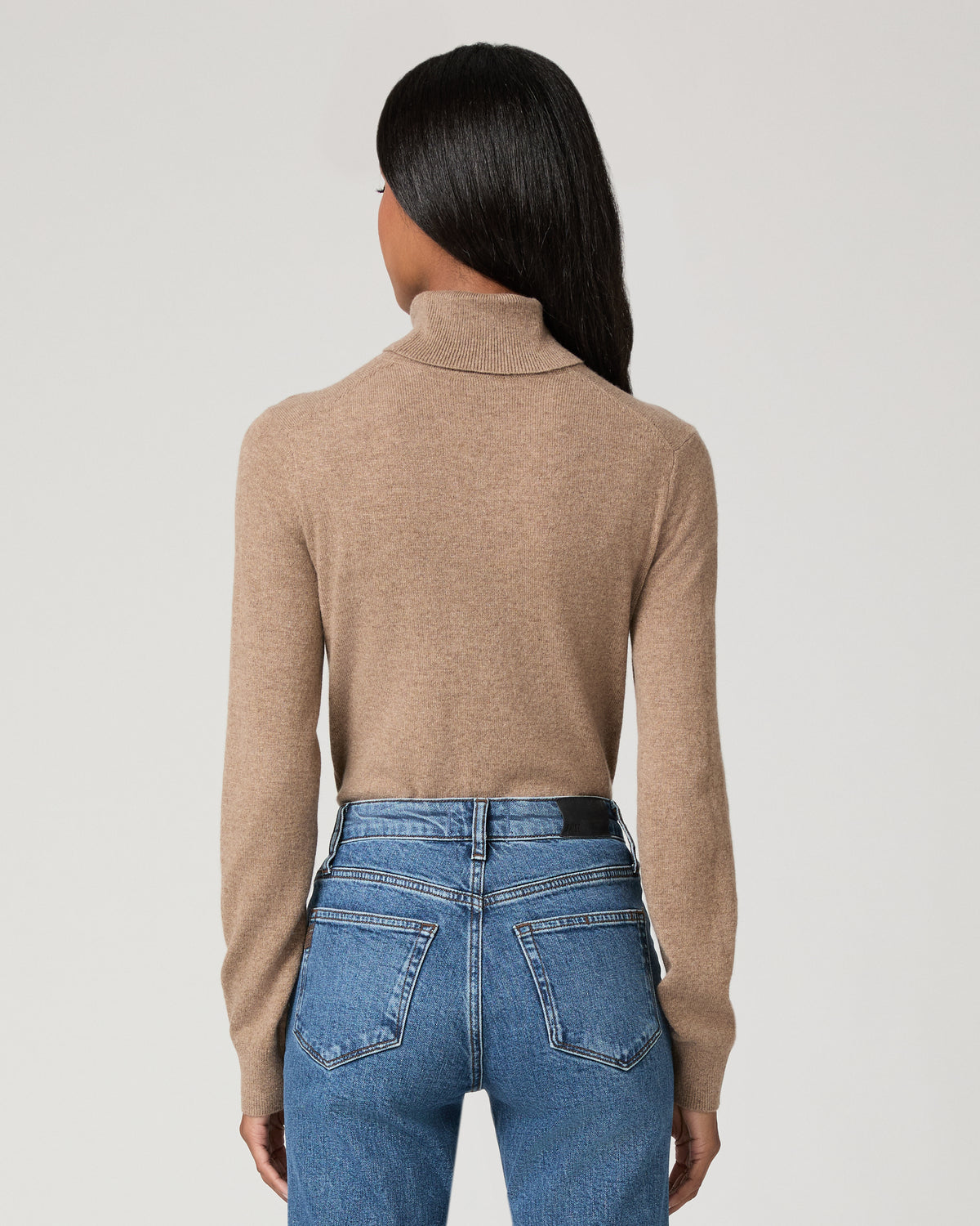 A model stands against a light gray studio background wearing a beige ribbed turtleneck sweater and blue high-waisted flare jeans with decorative silver buttons down the outer leg seams.