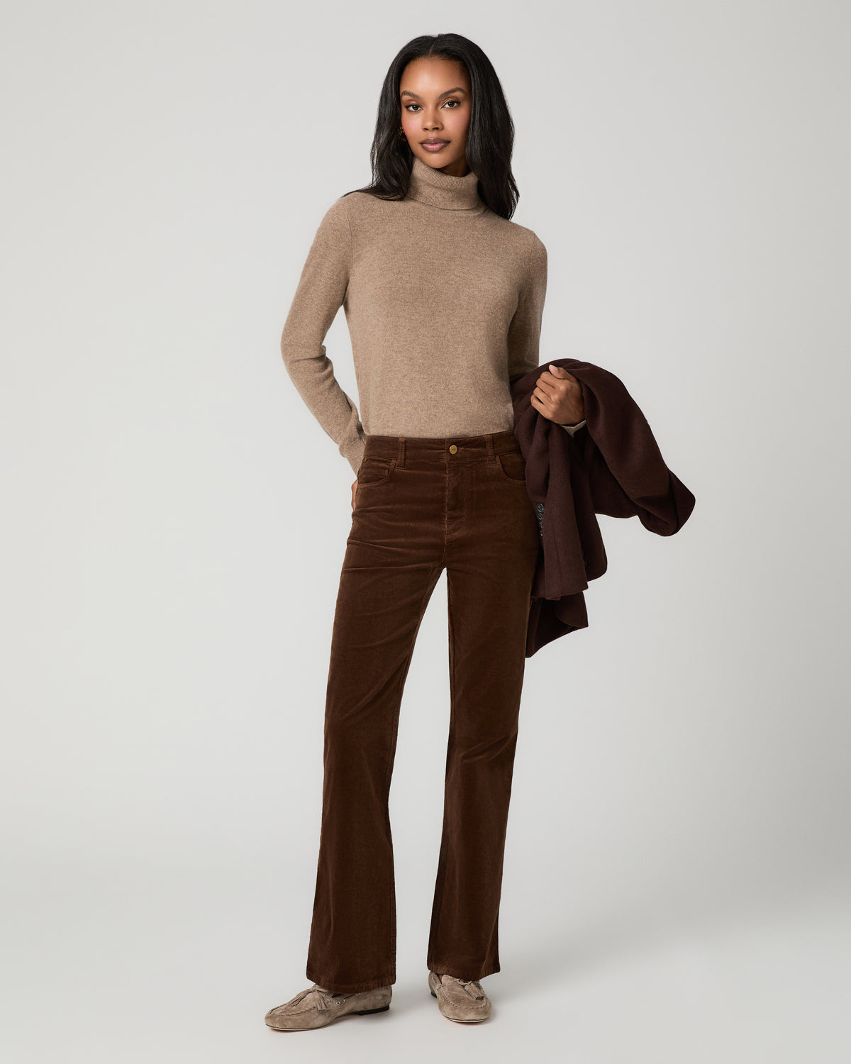 A model stands against a light gray studio background wearing a beige ribbed turtleneck sweater and blue high-waisted flare jeans with decorative silver buttons down the outer leg seams.