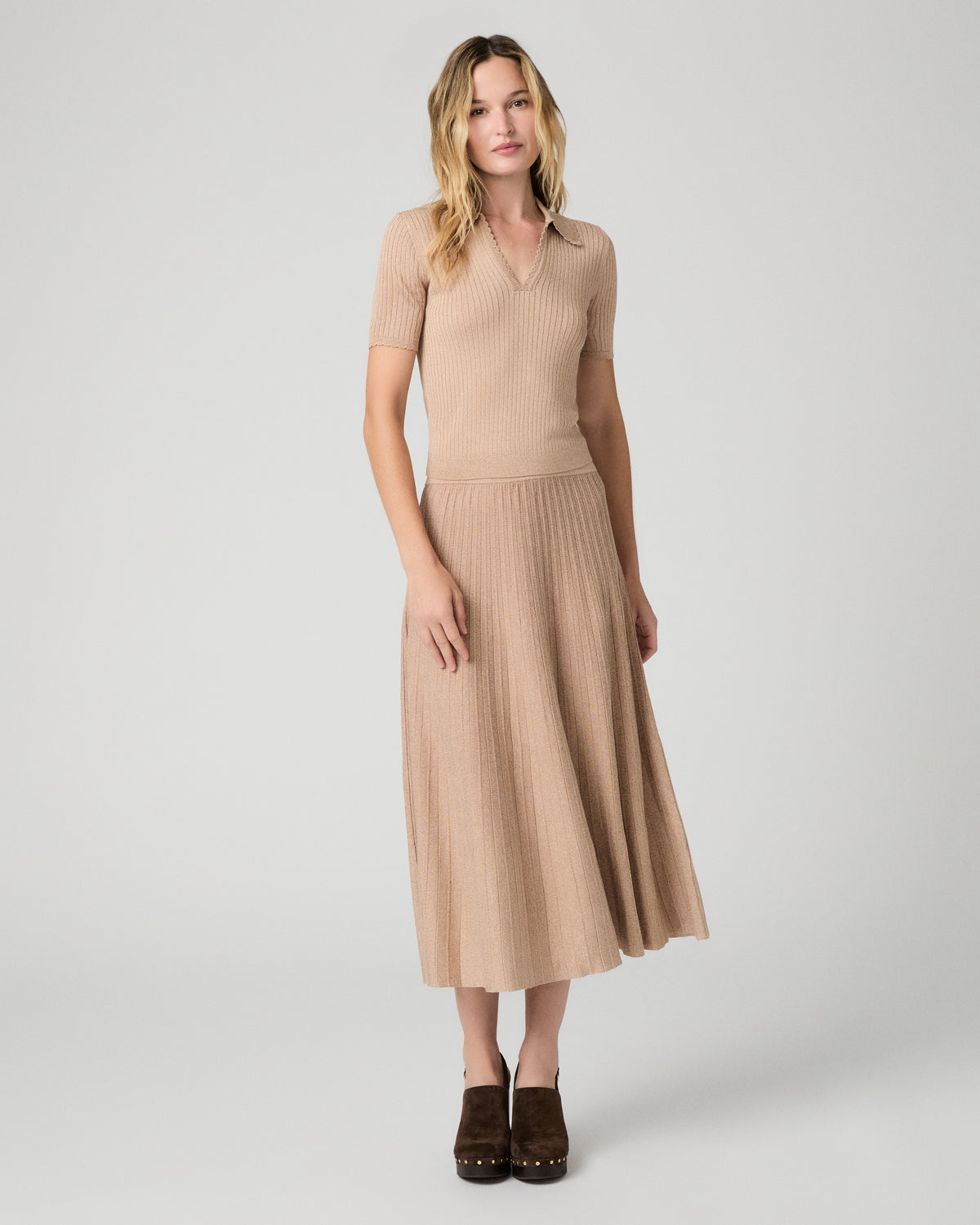 A woman modeling a beige ribbed knit midi dress with short sleeves, a V-neckline, and a pleated skirt, paired with dark brown shoes, against a light gray studio background.