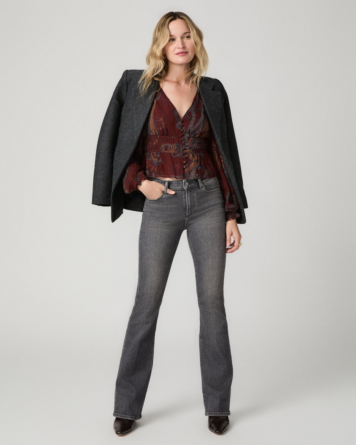 Full-length view of a woman wearing the garnet paisley-print Juna Blouse under an open black blazer, paired with gray flared jeans and black heels, one hand in her pocket.