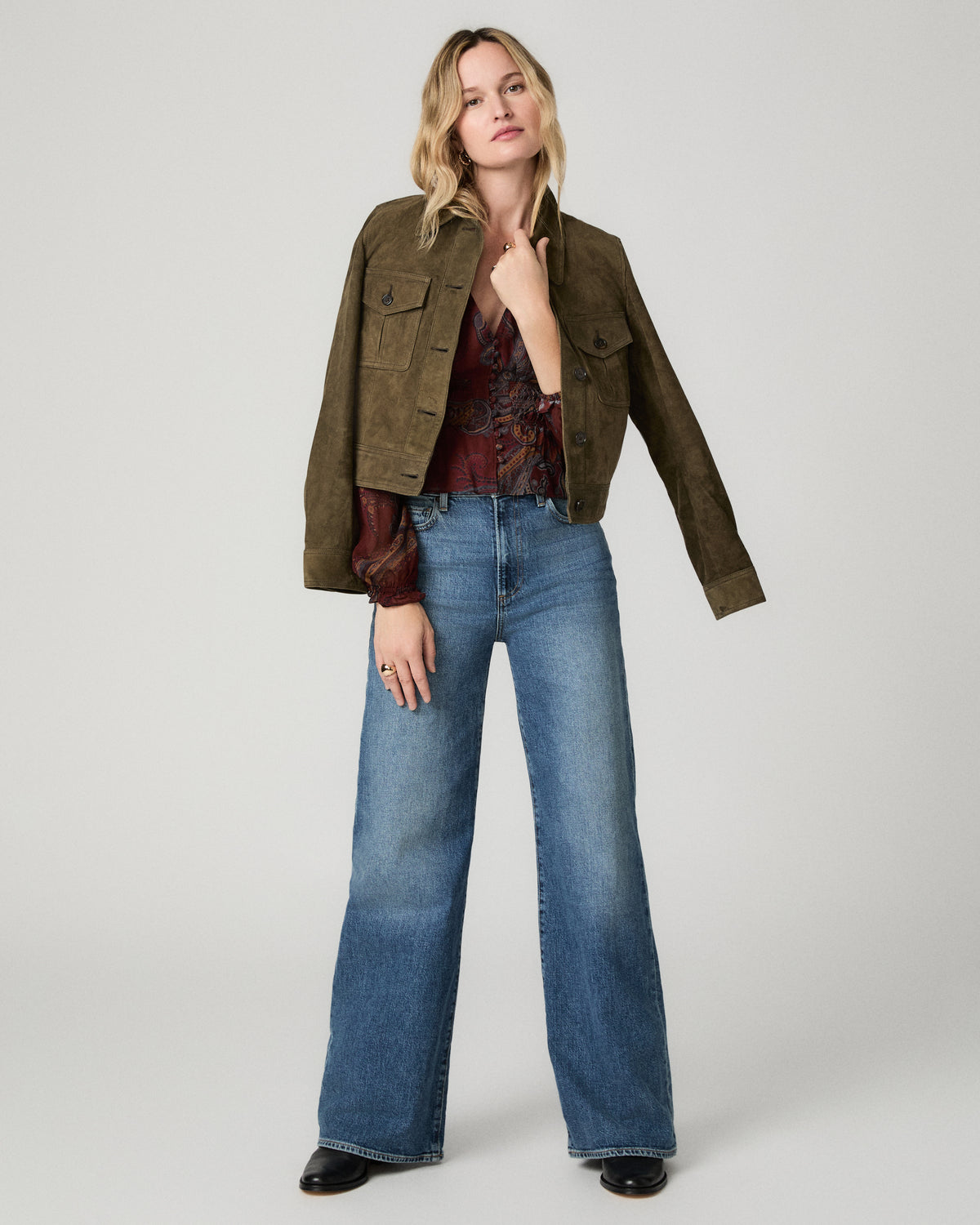 Full-length view of a woman wearing the garnet paisley-print Juna Blouse under an open olive green jacket, paired with wide-leg medium-wash jeans and black heels.