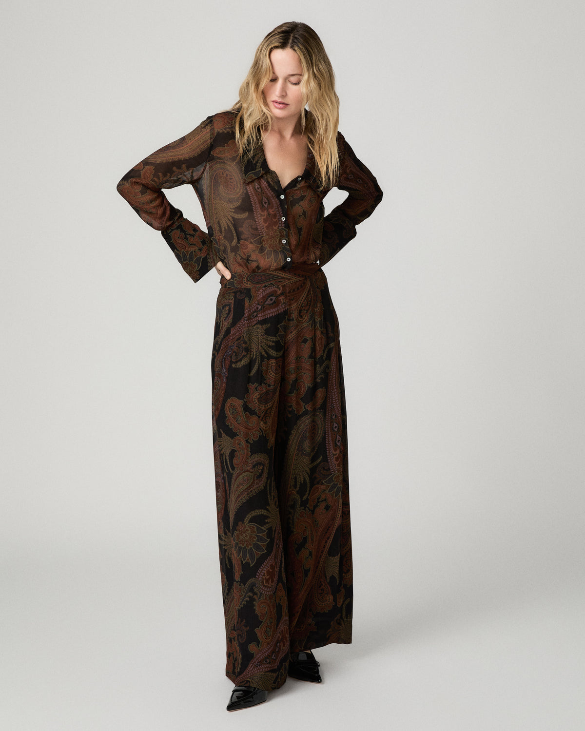 Full-length front view of a woman wearing the black and brown paisley-print Carina Pant with a matching paisley blouse, one hand on her hip.