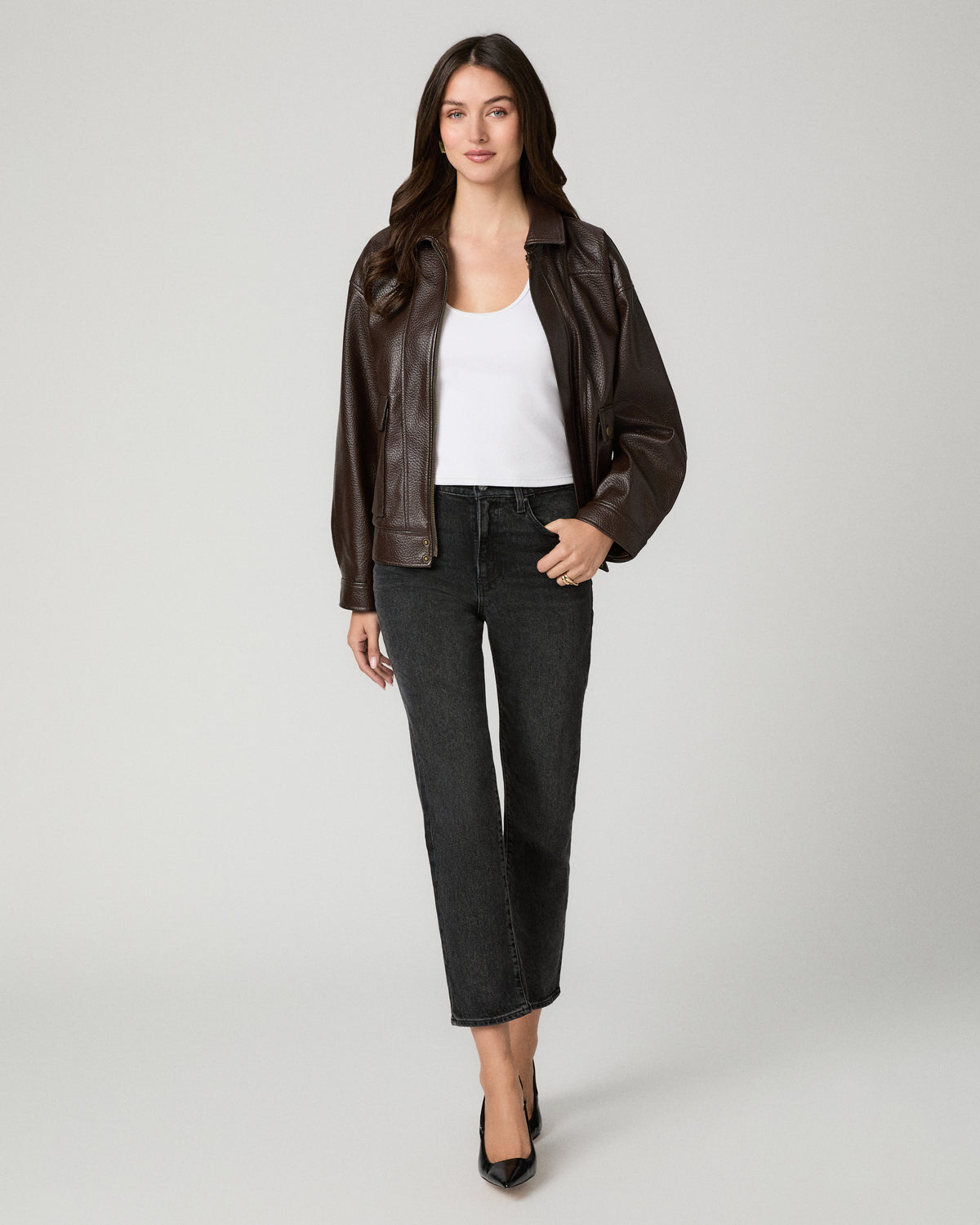 A woman modeling a dark brown faux-leather moto jacket over a white tank top and dark gray cropped jeans, standing against a light gray studio background.