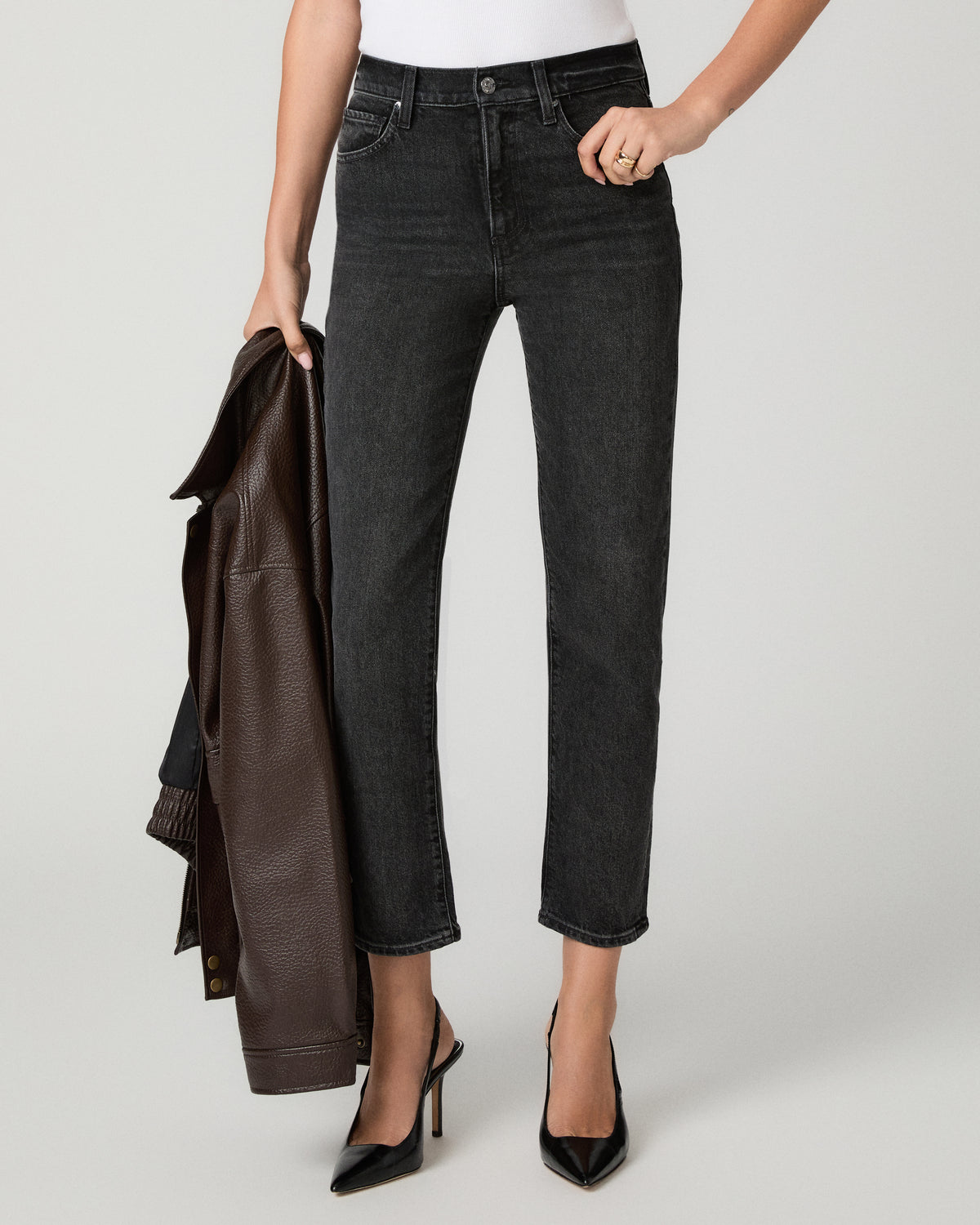 A model wearing dark gray cropped denim jeans, styled with a white top, black pointed-toe heels, and holding a brown leather jacket against a light gray studio background.