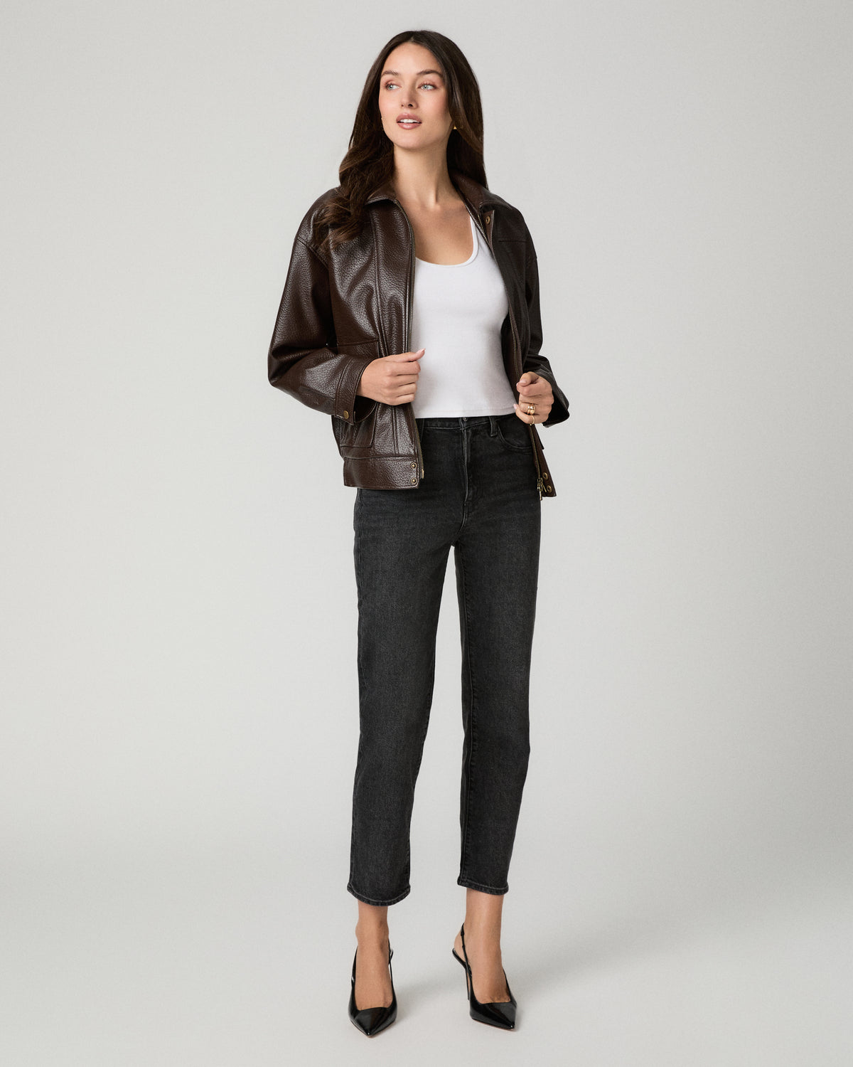 A woman modeling a dark brown faux-leather moto jacket over a white tank top, black cropped jeans, and black heels, photographed against a neutral light gray studio background.