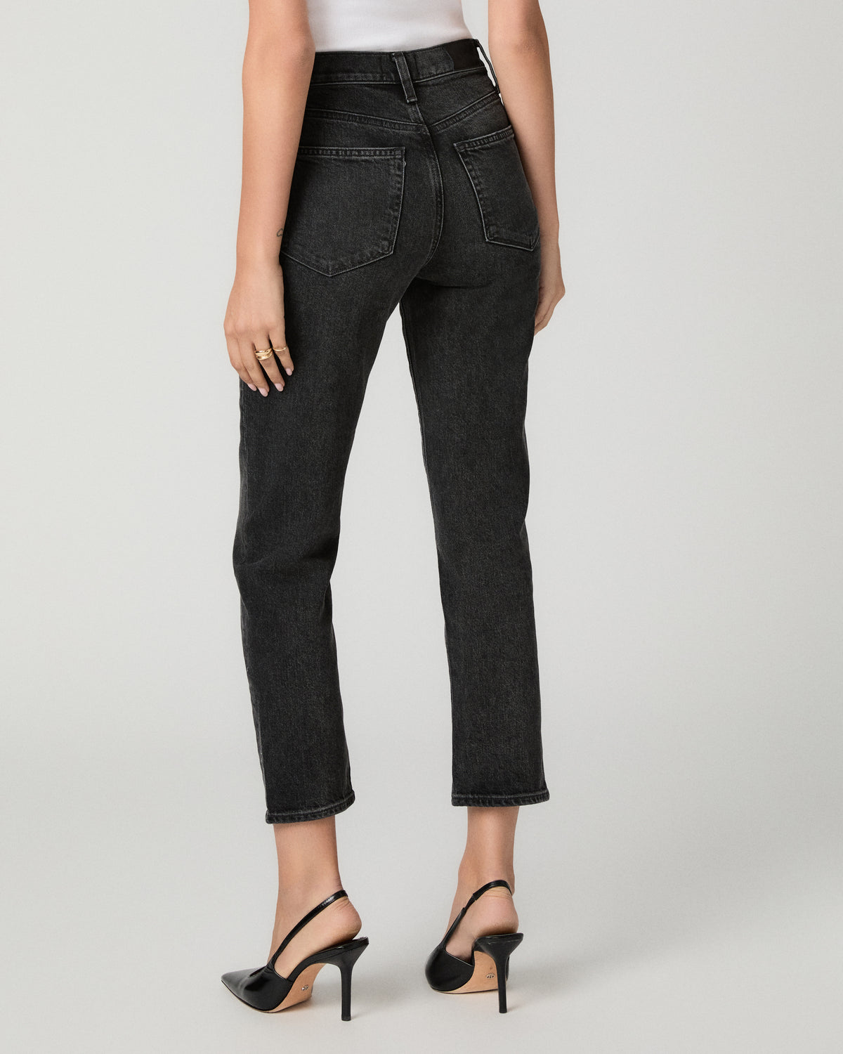 Rear view of a model wearing black cropped denim jeans with a straight-to-slightly-relaxed leg, paired with black slingback heels against a light gray studio background.