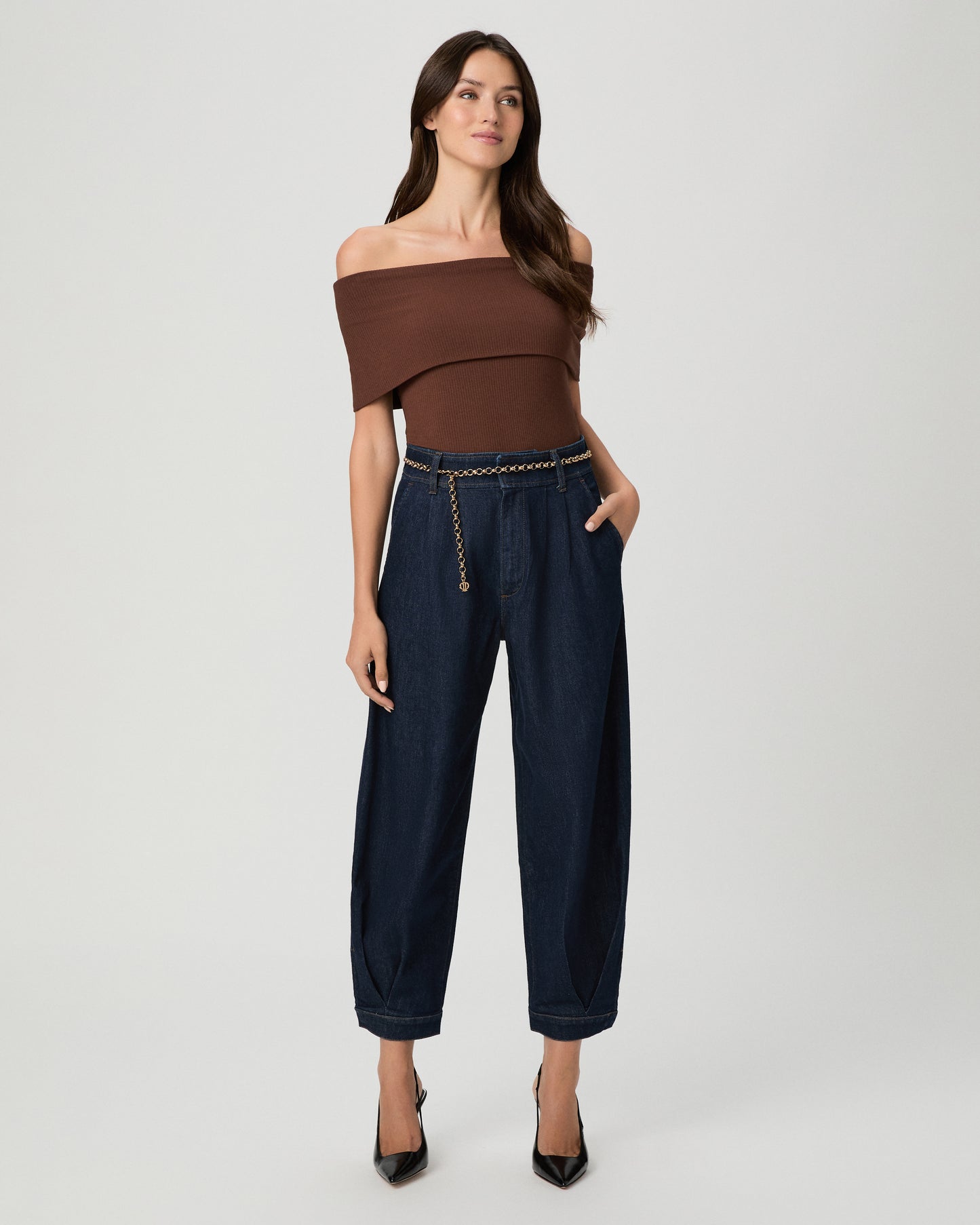 A woman wearing a brown off the shoulder top paired with dark denim pants with a gold belt and black heels against a light gray studio background
