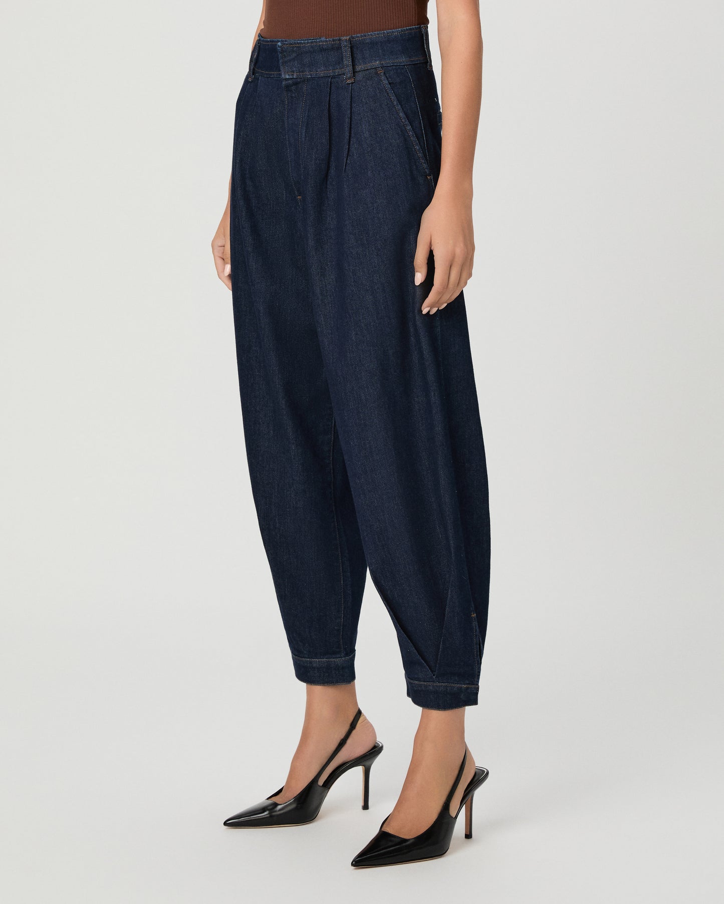 A woman wearing a brown off the shoulder top paired with dark denim pants with black heels against a light gray studio background