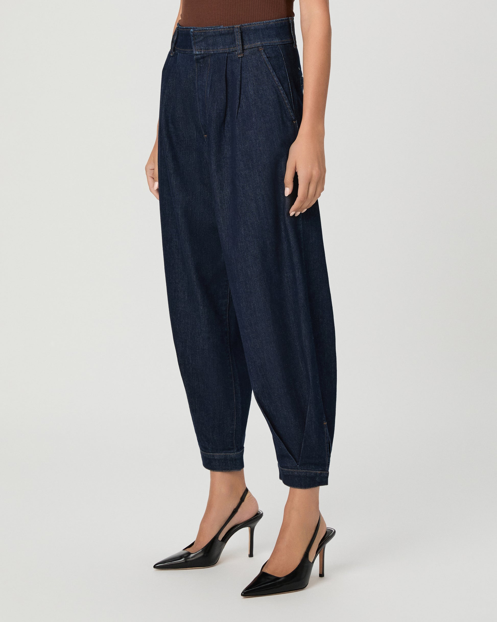 A woman wearing a brown off the shoulder top paired with dark denim pants with black heels against a light gray studio background