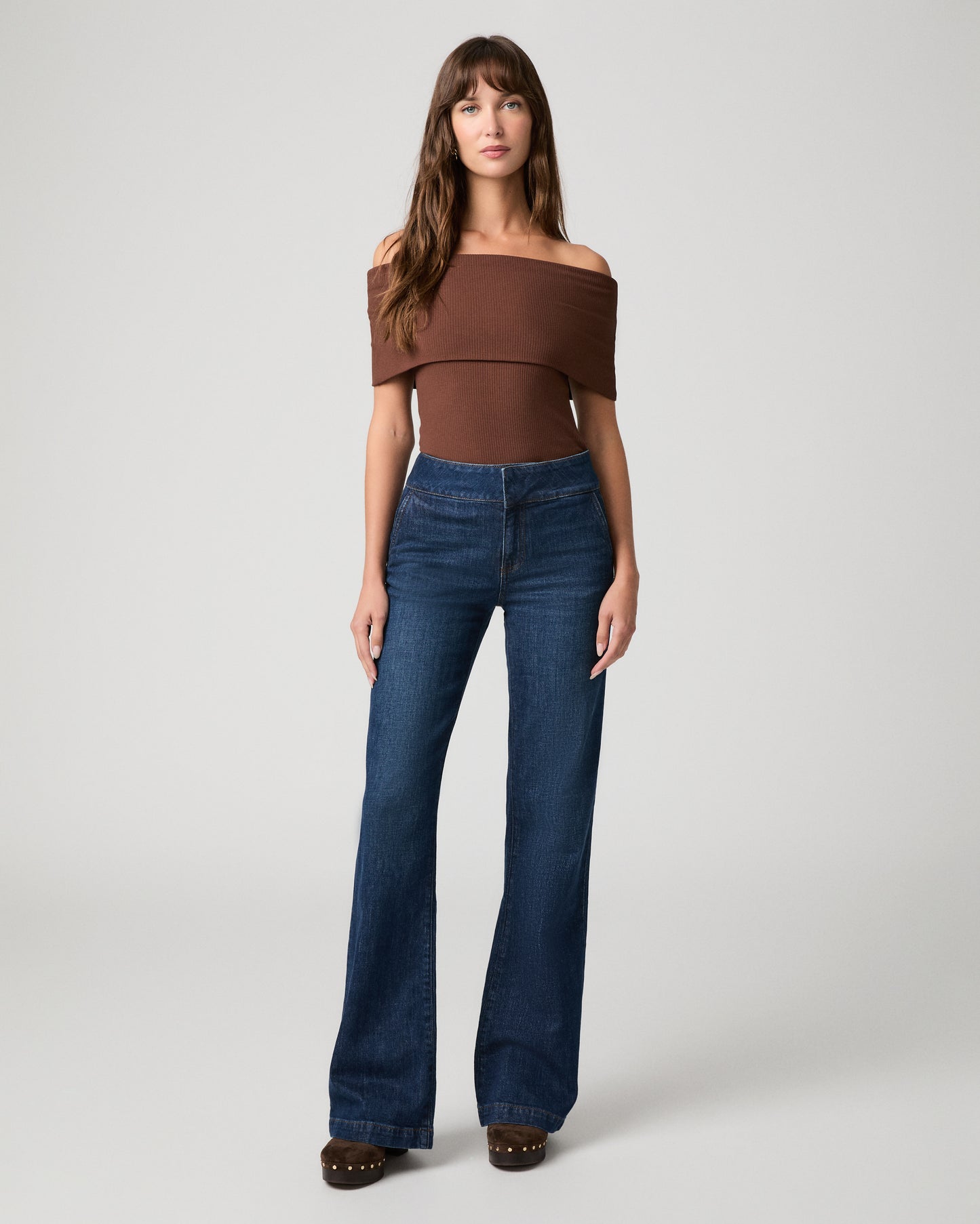Woman wearing brown top and blue pants with a white background.
