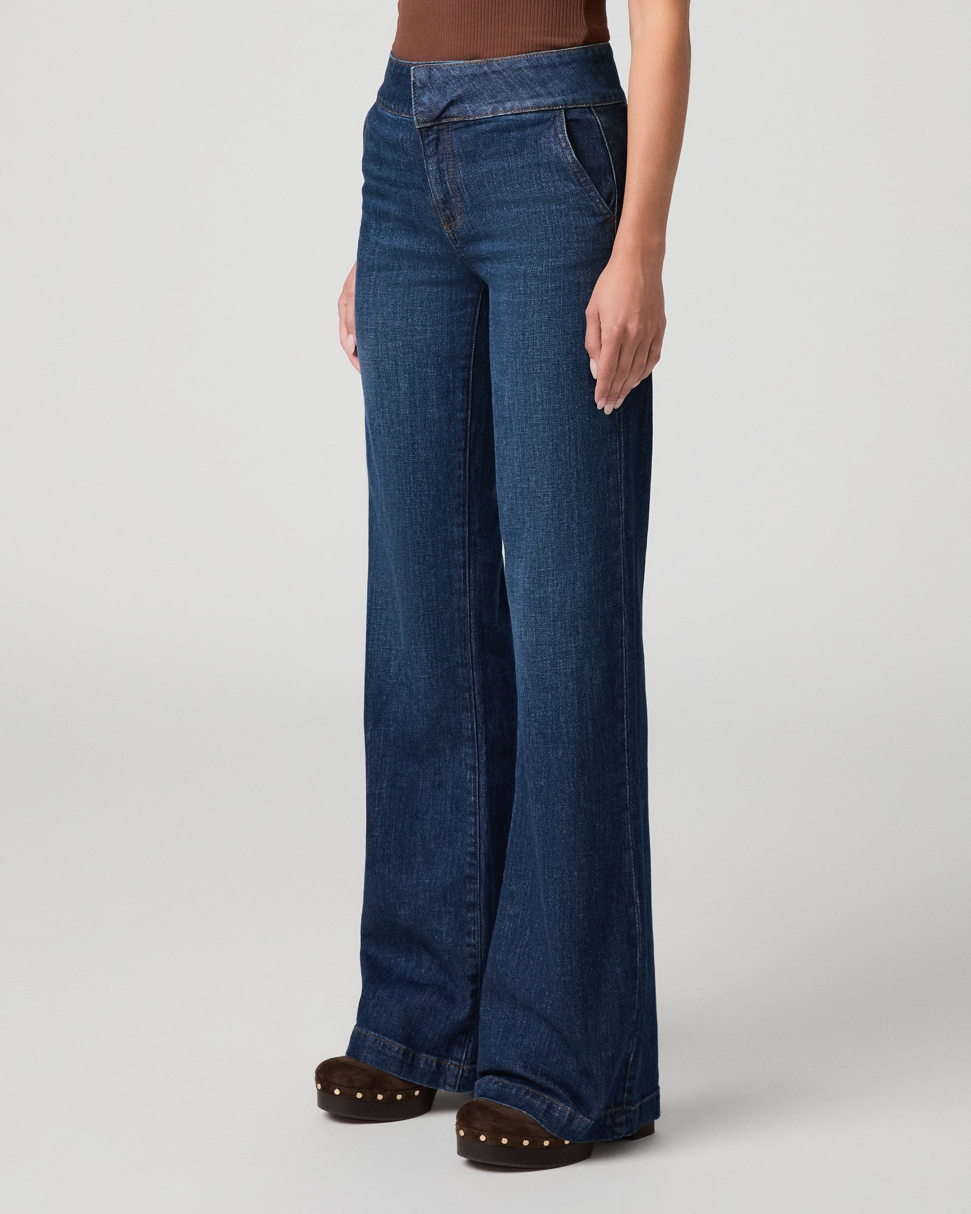 Woman wearing brown top and blue pants with a white background.