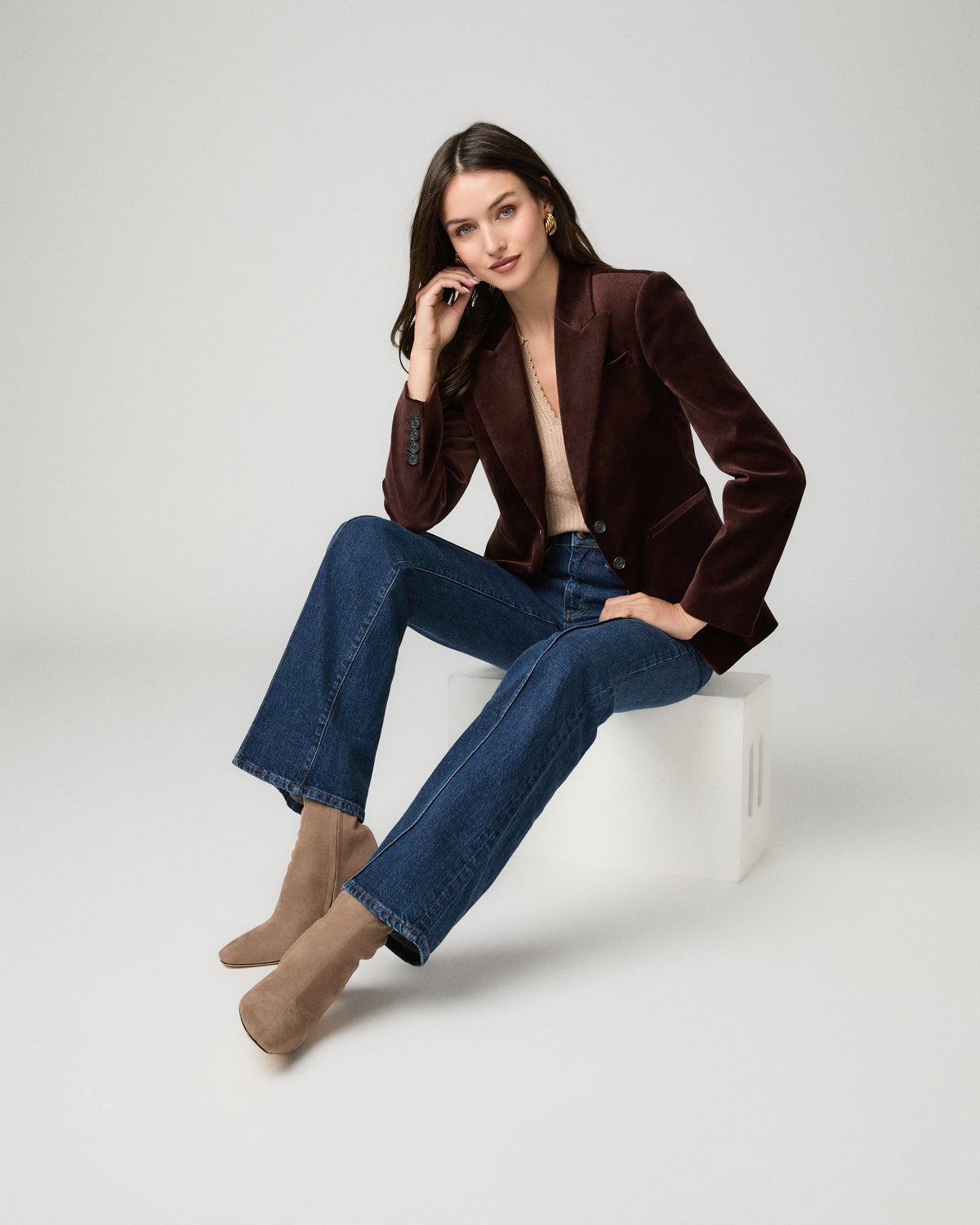 A woman models a dark brown velvet blazer paired with blue jeans and beige ankle boots, sitting on a white block against a light gray studio background.