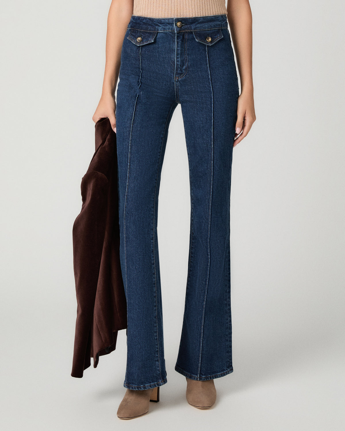 A model wearing dark blue high-waisted flare jeans, holding a dark brown coat, against a light gray studio background.