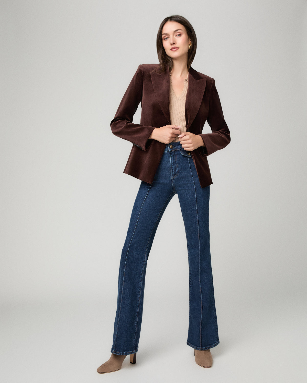 A woman modeling a dark brown velvet blazer with flared sleeves, paired with blue flare jeans and beige heels, against a light gray studio background.