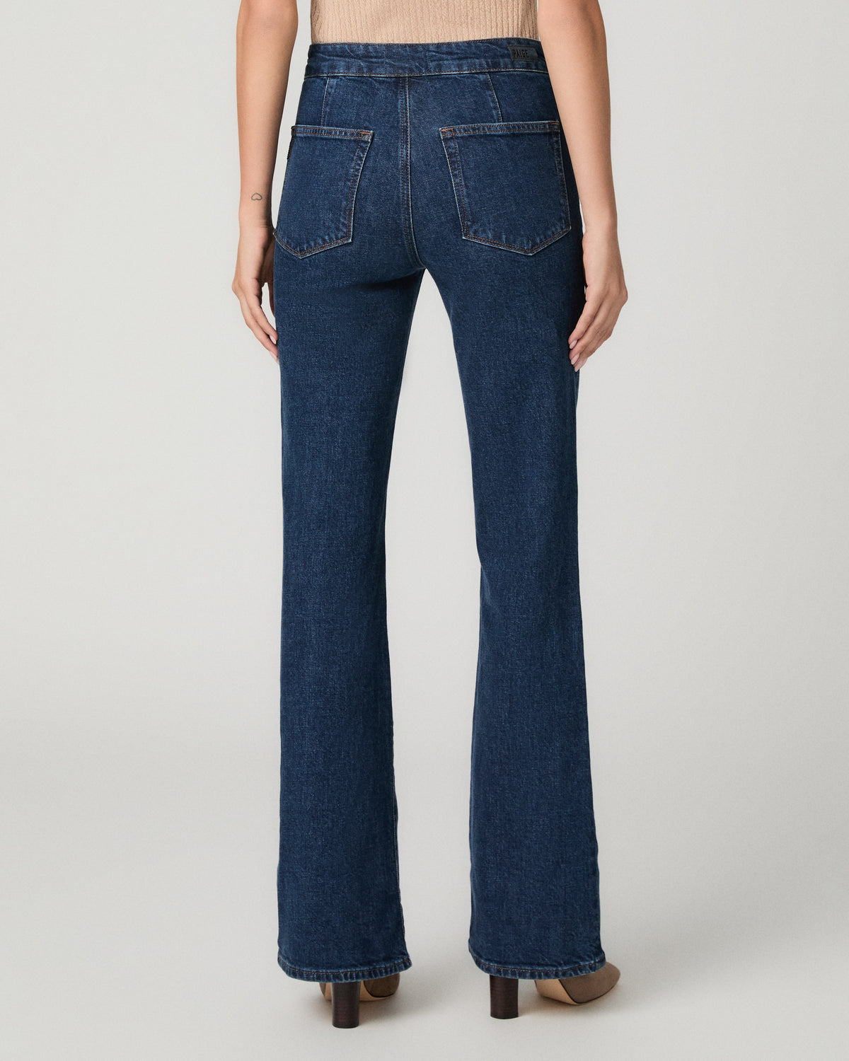 A woman modeling a tan top, paired with blue flare jeans and beige heels, against a light gray studio background.