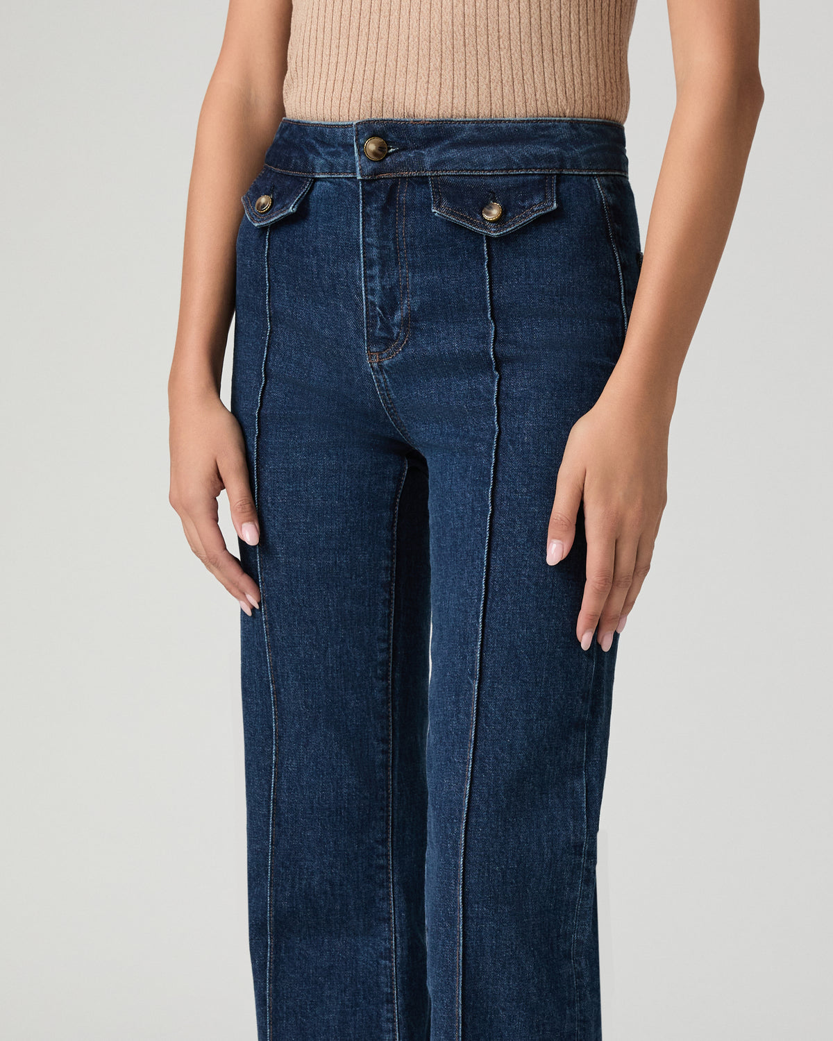 A model wearing dark blue high-waisted straight-leg jeans with front flap pockets and gold-tone buttons, paired with a beige ribbed knit top against a light gray studio background.