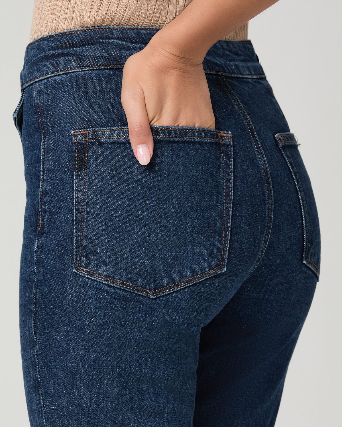 Close-up back view of a person wearing dark blue denim jeans, showing the back pocket with a hand inserted, against a light gray studio background.